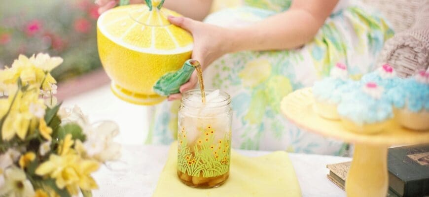 A yellow picnic display with a woman holding a ceramic lemon-shaped pot, a cake stand with cupcakes, a vase with flowers, and a jar of iced tea sitting on a yellow board